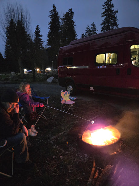 Family camping at dusk: A child roasts marshmallows by the campfire with an adult, next to their red RV rental camper van.