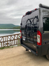 Dark gray Rize camper van with a bike rack by a scenic lake and mountains, perfect for an RV rental outdoor adventure.