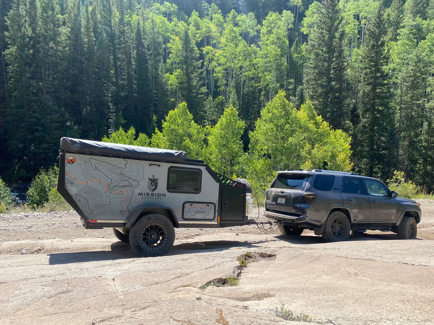 This rugged overland RV rental, a trailer towed by an SUV, is set for adventure and off-grid camping amidst a sunny forest.