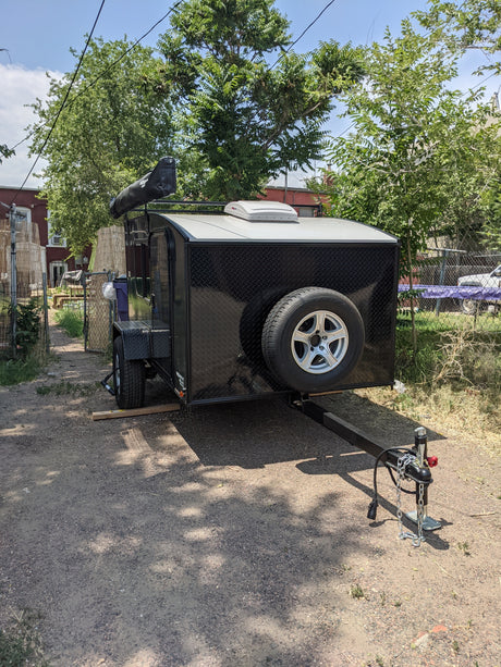 Compact black RV rental trailer with awning, spare tire, and roof vent, parked on a gravel path.
