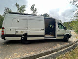 Experience the open road in this white Mercedes Sprinter rv rental camper van with open side door revealing interior. Equipped with external hookups & roof rack.