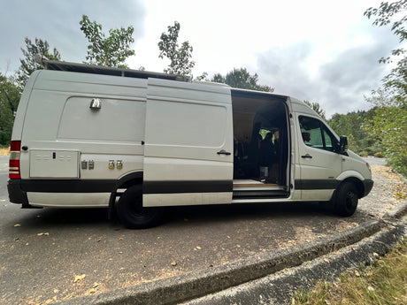 Experience the open road in this white Mercedes Sprinter rv rental camper van with open side door revealing interior. Equipped with external hookups & roof rack.