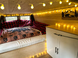 Cozy RV rental interior featuring warm string lights, a comfortable bed with bear print blanket, and custom wood paneling.