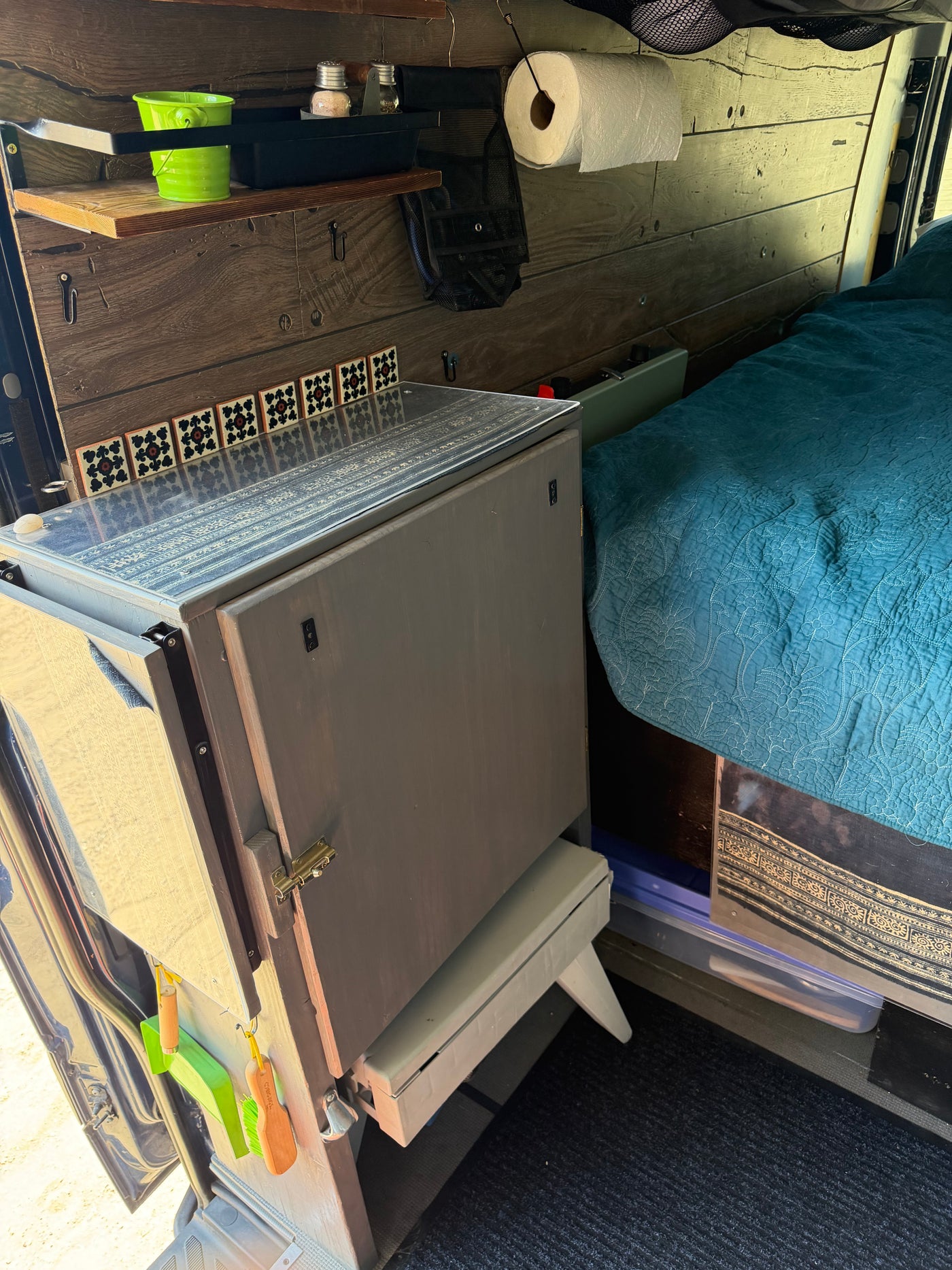 Cozy RV rental interior with wood-paneled walls, compact kitchenette, shelves, and a teal quilted bed. A pull-out step is ready for use. — photo 9