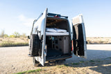 This RV rental's open rear reveals a custom camper van interior with a cozy bed, perfect for a road trip adventure.