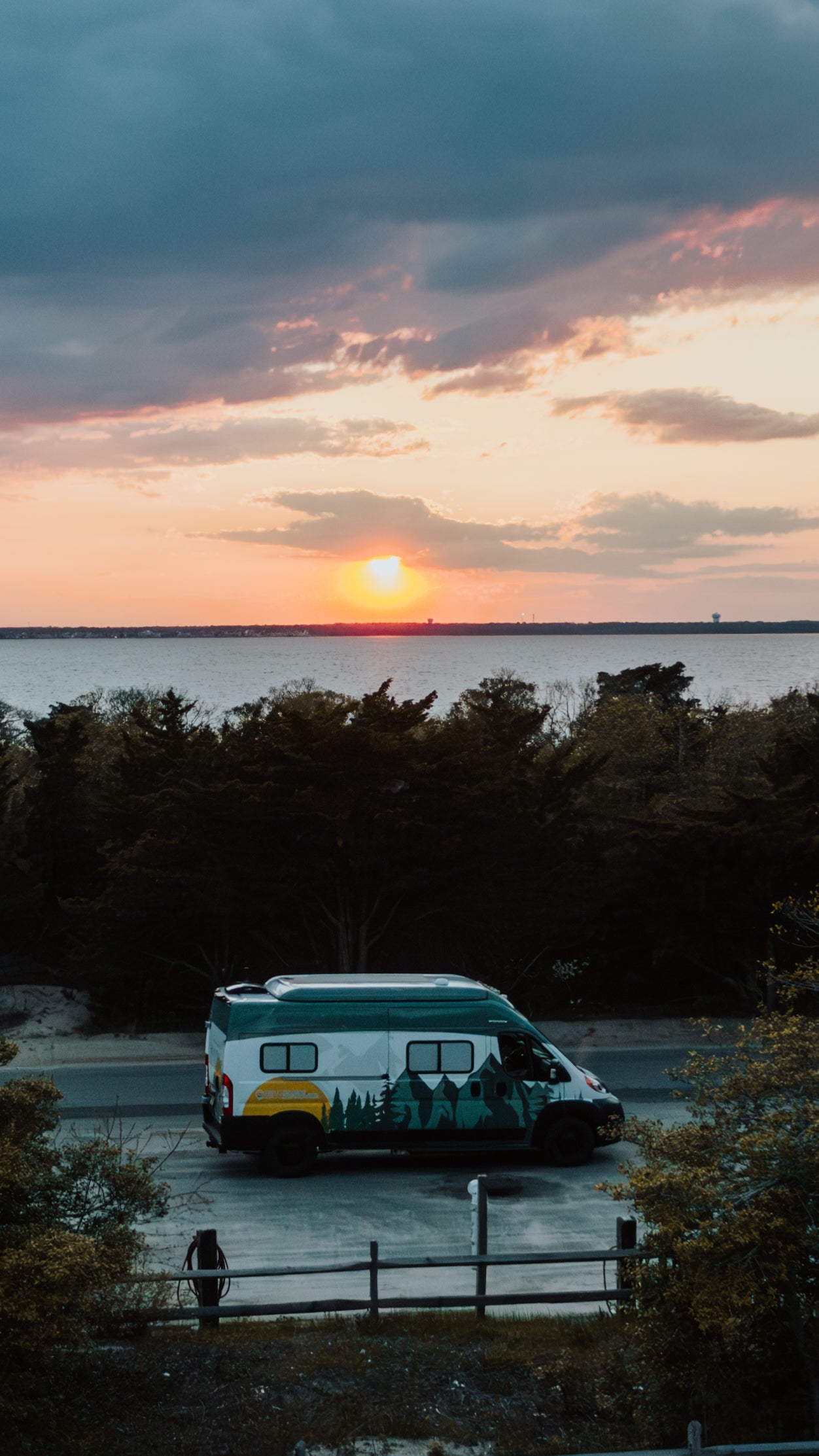 RV rental: A custom camper van with mountain graphics, parked at a scenic sunset by a calm bay.