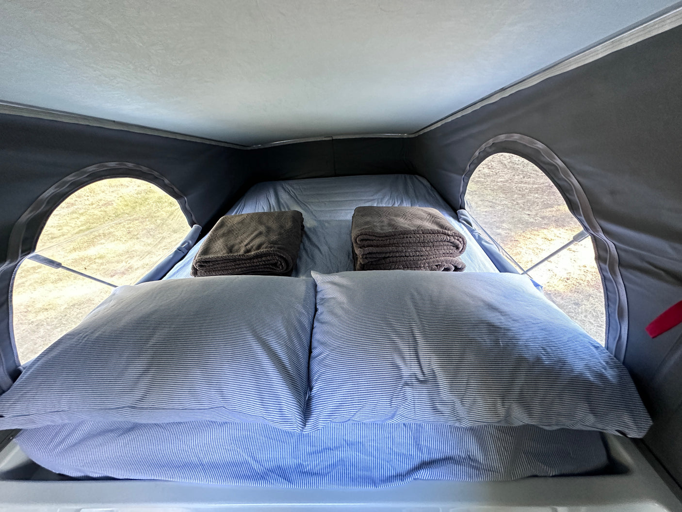 Cozy RV rental sleeping area with striped pillows and folded blankets, through pop-top windows see green outside.