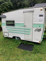 A charming vintage Shasta Compact travel trailer, in white and green, sits on grass, ready for an RV rental.
