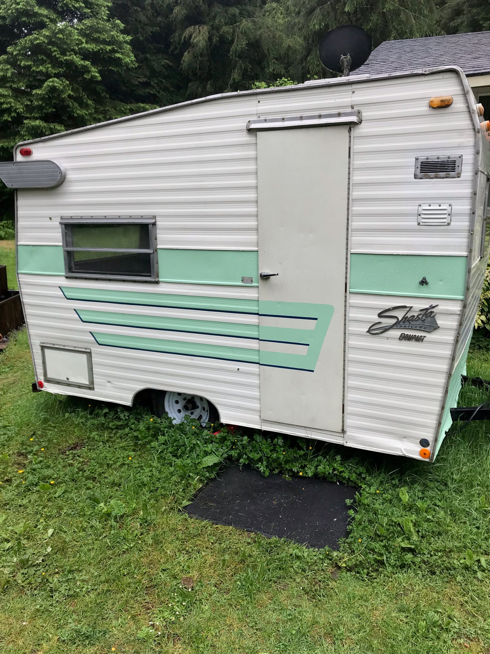 A charming vintage Shasta Compact travel trailer, in white and green, sits on grass, ready for an RV rental.