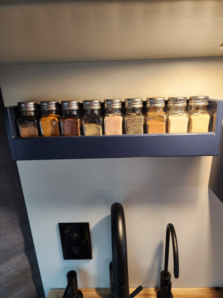 Compact spice rack in an RV rental kitchen, showing labeled spice jars next to a black faucet and switch.