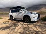 Rugged white Lexus GX SUV with a rooftop tent parked on sand near mountains, ideal for an adventure or RV rental trip.