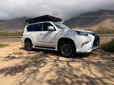 Rugged white Lexus GX SUV with a rooftop tent parked on sand near mountains, ideal for an adventure or RV rental trip.