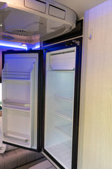 Inside an rv rental: open mini-fridge and freezer, AC unit above, illuminated by blue light.