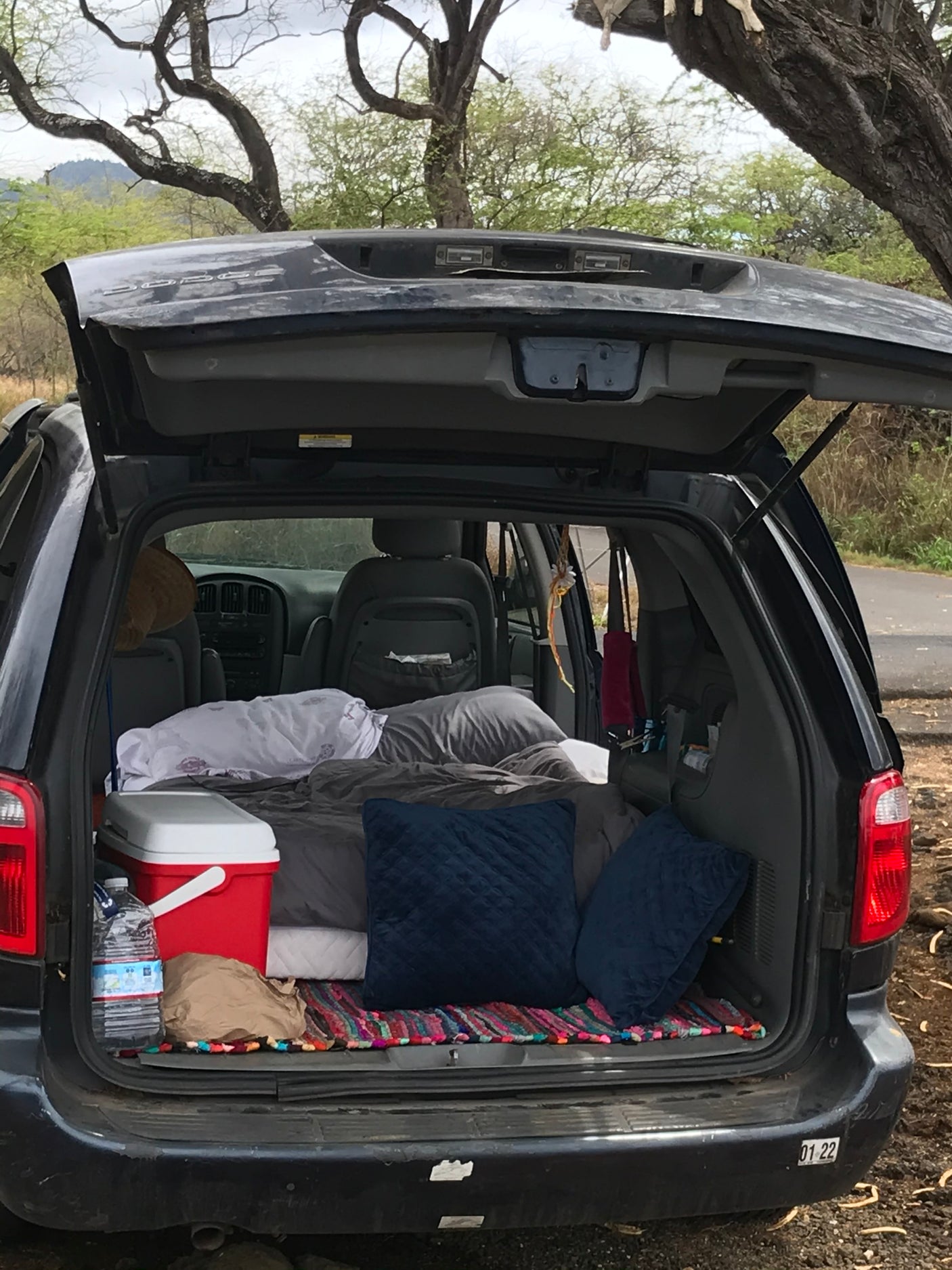 Rear of a Dodge minivan opened for car camping. Features a cozy sleep setup with blankets, pillows, and a cooler. An RV rental alternative.