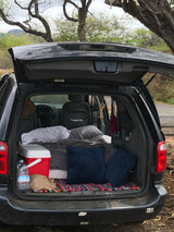 Rear of a Dodge minivan opened for car camping. Features a cozy sleep setup with blankets, pillows, and a cooler. An RV rental alternative.