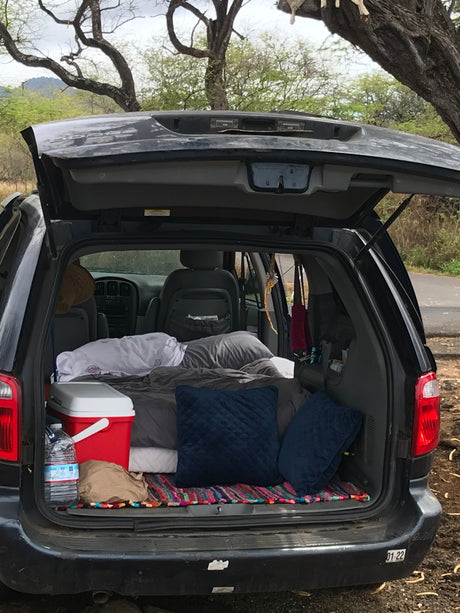 Rear of a Dodge minivan opened for car camping. Features a cozy sleep setup with blankets, pillows, and a cooler. An RV rental alternative.
