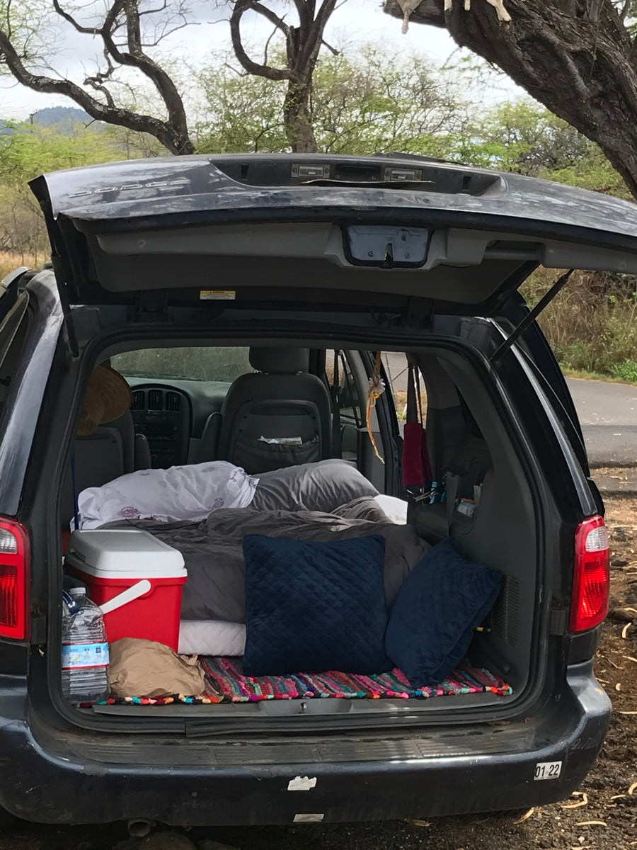 Rear of a Dodge minivan opened for car camping. Features a cozy sleep setup with blankets, pillows, and a cooler. An RV rental alternative.