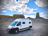 White RV rental parked in a vast desert landscape with striking rock formations under a cloudy blue sky, suggesting adventure.