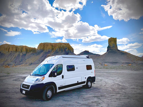White RV rental parked in a vast desert landscape with striking rock formations under a cloudy blue sky, suggesting adventure.