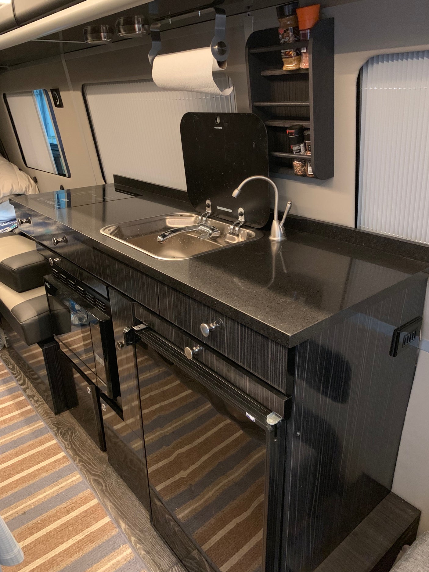 A sleek RV rental kitchen features a dark counter with a sink, stovetop, under-counter appliances, and overhead spice rack.