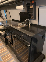 A sleek RV rental kitchen features a dark counter with a sink, stovetop, under-counter appliances, and overhead spice rack.