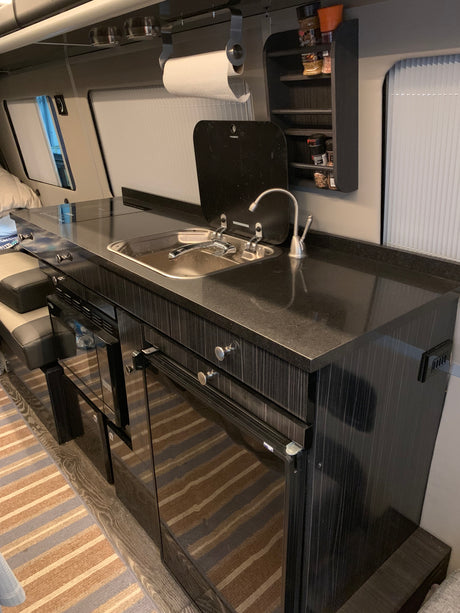 A sleek RV rental kitchen features a dark counter with a sink, stovetop, under-counter appliances, and overhead spice rack.