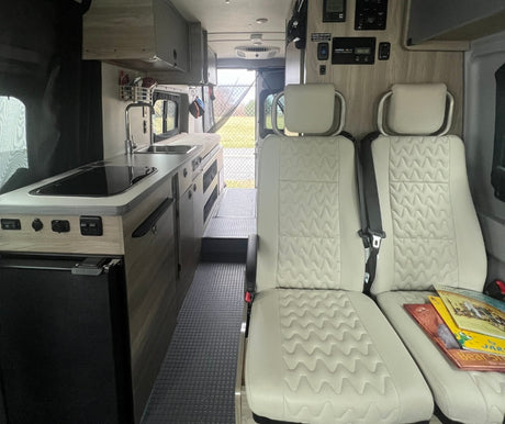 Clean RV rental interior featuring a functional kitchenette with a sink and stovetop, plus two comfortable passenger seats.