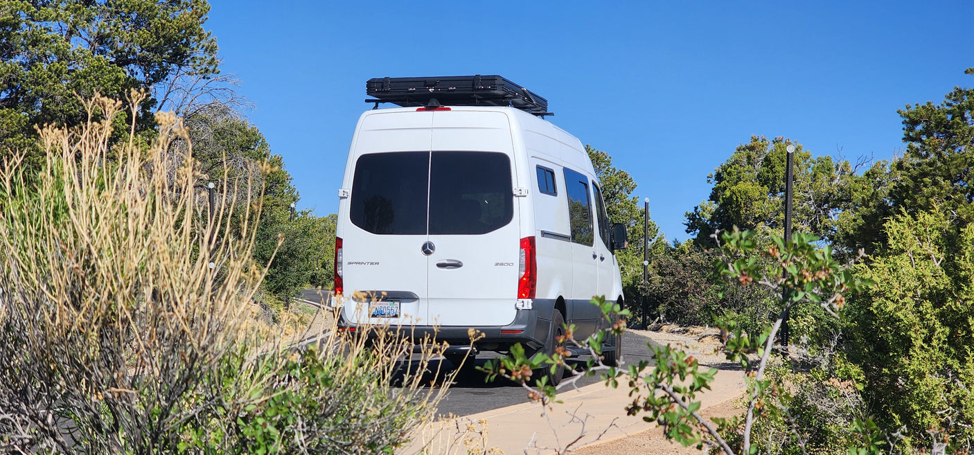White Mercedes Sprinter camper van, likely an RV rental, on a scenic road with dry brush and green trees under clear skies.