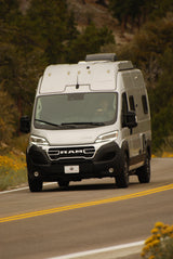 A silver Winnebago Ram ProMaster camper van RV rental drives on a scenic road with trees, ready for adventure.