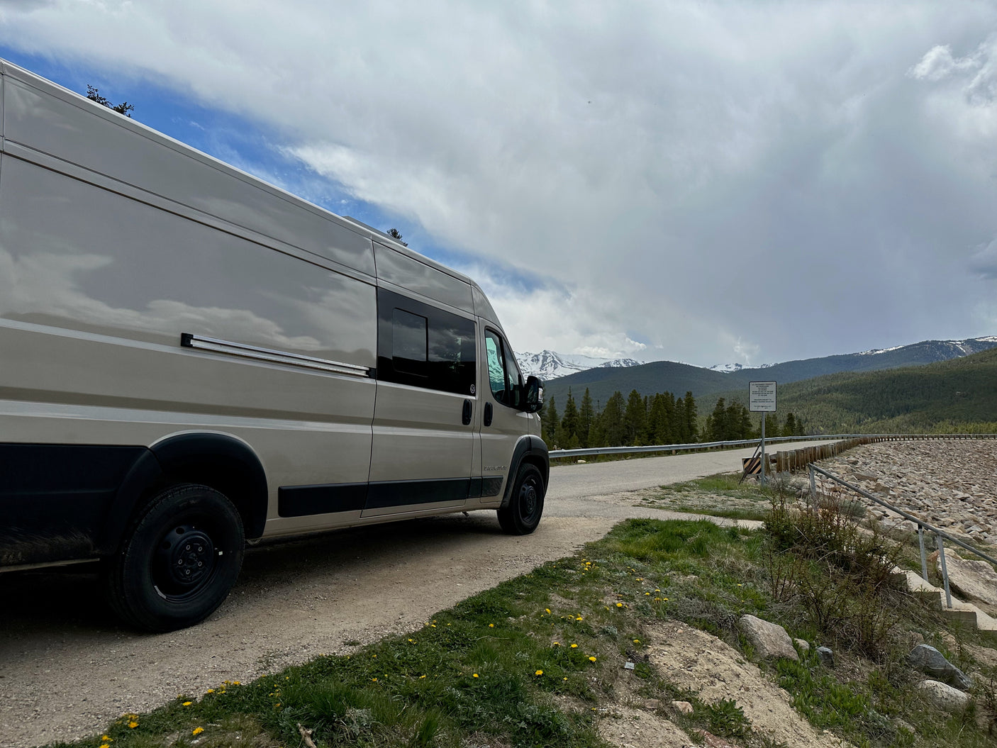 Beige rv rental van on a dirt road with yellow flowers. Snowy mountains, green forest, and cloudy sky in background.