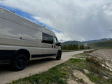 Beige rv rental van on a dirt road with yellow flowers. Snowy mountains, green forest, and cloudy sky in background.