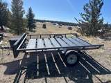 Black utility trailer in a sunny mountain landscape with pines & homes. Great for gear with an RV rental.