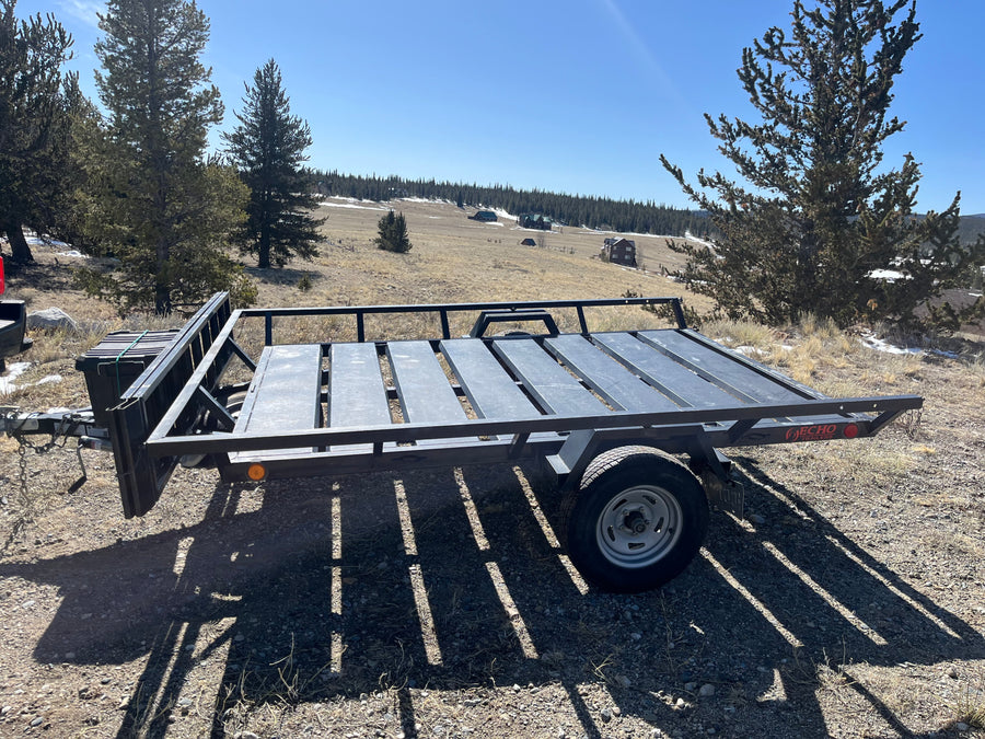 Black utility trailer in a sunny mountain landscape with pines & homes. Great for gear with an RV rental.