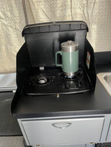 RV rental: Compact kitchen with a Suburban two-burner stove and sink. A green insulated mug is ready on the burner.