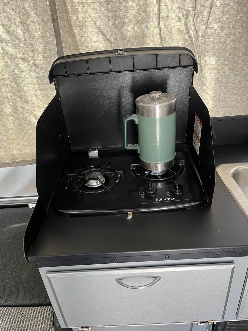 RV rental: Compact kitchen with a Suburban two-burner stove and sink. A green insulated mug is ready on the burner. — photo 14