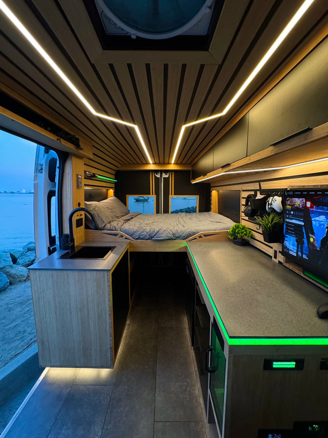 Modern custom RV rental interior with wooden ceiling, LED lighting, cozy bed, kitchen sink, and a cool gaming setup by the water.