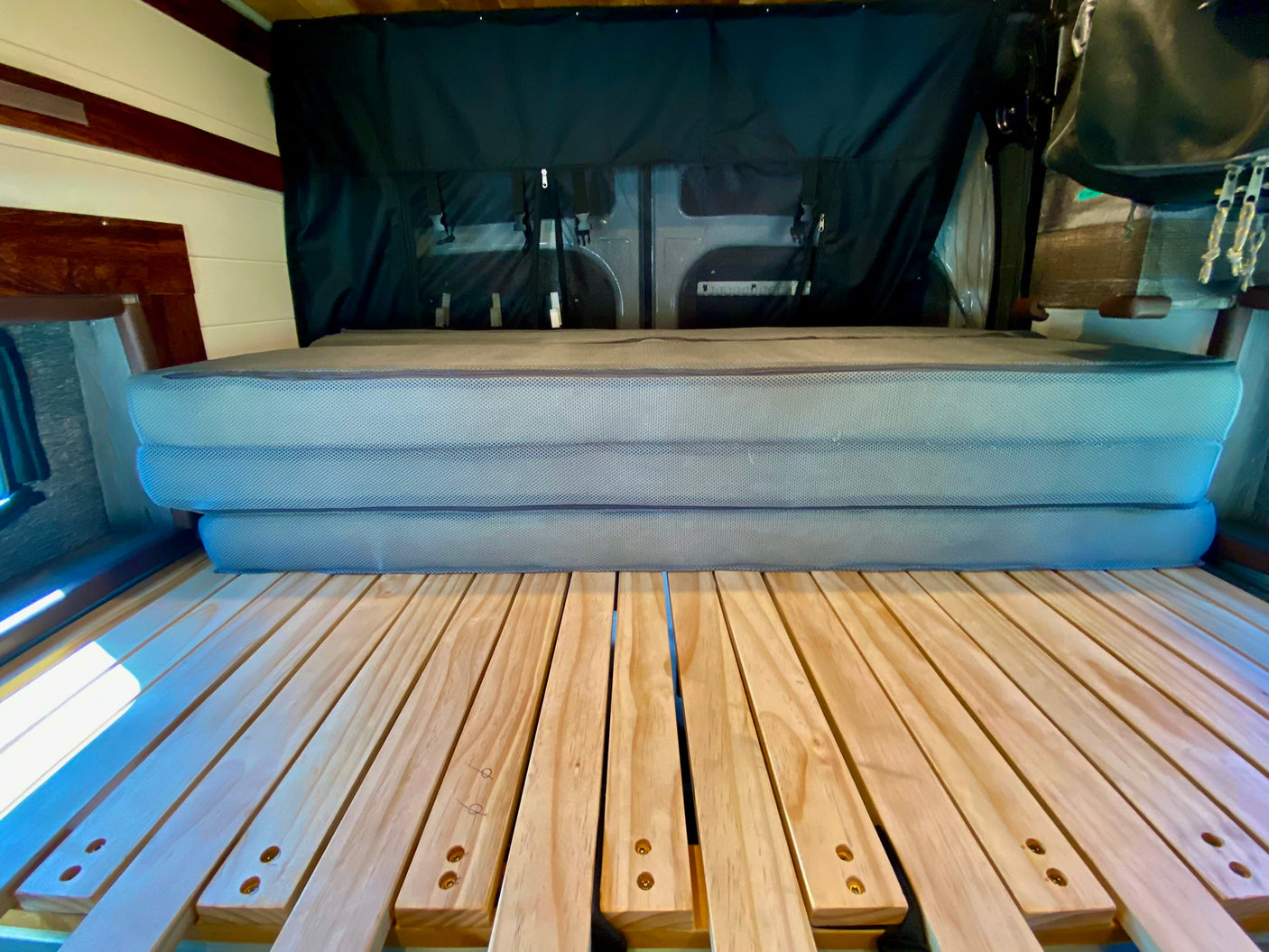 RV rental interior: Wooden slat bed frame, stacked grey mattresses, & rear storage curtain in a camper van.