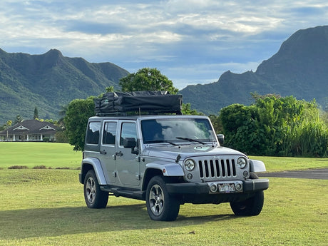 This silver Jeep Wrangler with a rooftop tent, like an RV rental, is parked on a green field with Hawaiian mountains behind.