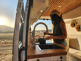 RV rental cooking with a view! A person stirs food in a campervan kitchen, open to golden hills at sunrise/sunset.