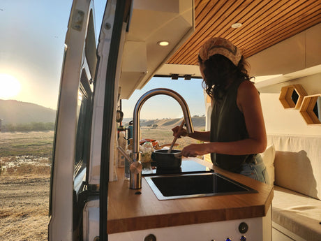 RV rental cooking with a view! A person stirs food in a campervan kitchen, open to golden hills at sunrise/sunset.