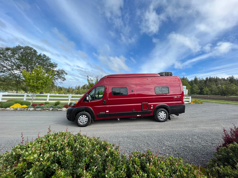 Red RV rental van parked on a gravel drive, with trees, a white fence, and blue sky overhead. — photo 1