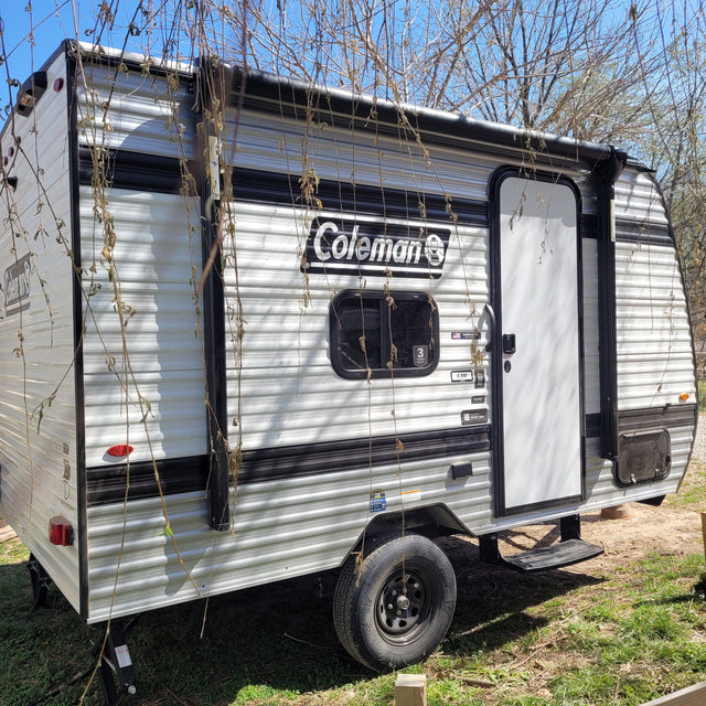 This Coleman travel trailer, ideal for an RV rental, is parked outdoors. It's a compact camper under leafless branches.