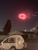Red fireworks explode over houses at night, with a sleek, teardrop-shaped RV rental parked in the foreground.