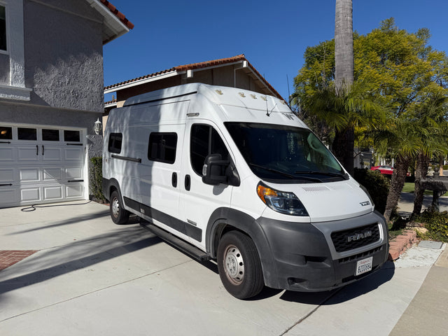 White Winnebago Solis RV rental camper van parked in a sunny driveway, plugged in for charging at home.