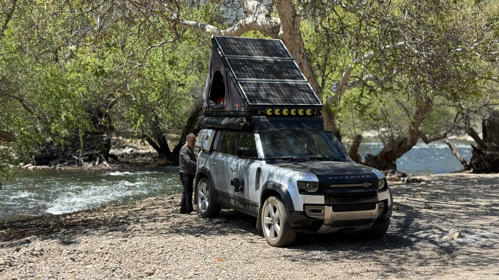 Adventure-ready Land Rover Defender with rooftop tent & solar panels by a river. An RV rental option for wilderness exploration.
