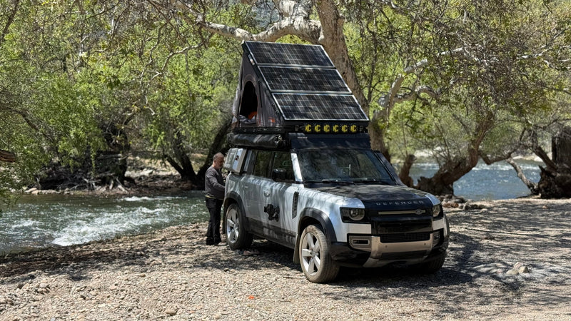 Adventure-ready Land Rover Defender with rooftop tent & solar panels by a river. An RV rental option for wilderness exploration. — photo 1