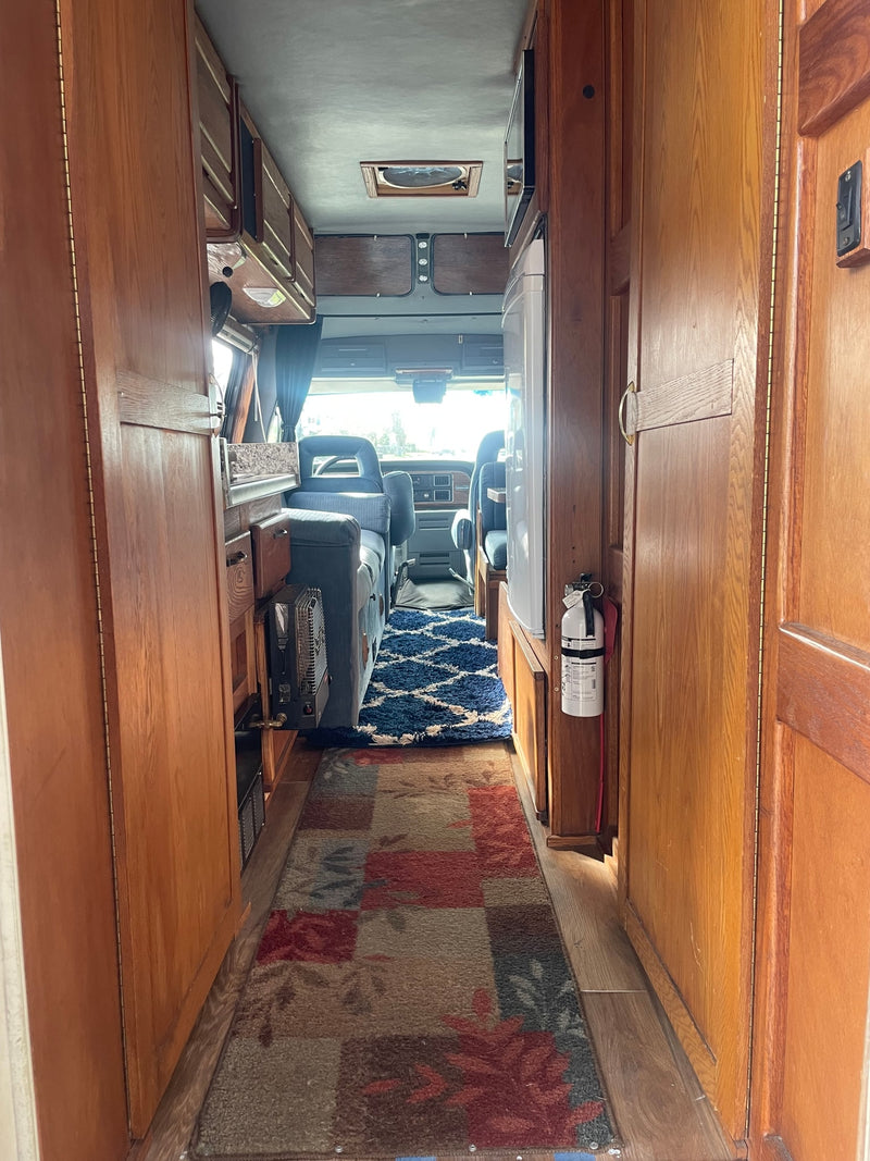 Cozy RV rental interior: wooden cabinetry, narrow aisle with rugs, microwave, fridge, and front seats visible. — photo 2