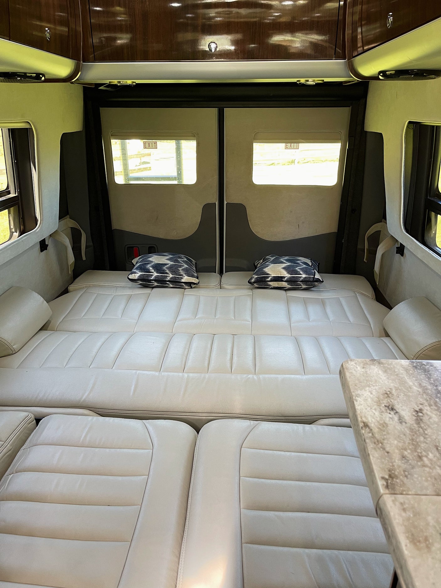 RV rental camper van interior showing light leather bed setup with patterned pillows, overhead cabinets, and windows displaying 'EXIT' signs.
