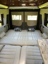 RV rental camper van interior showing light leather bed setup with patterned pillows, overhead cabinets, and windows displaying 'EXIT' signs.