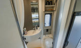 Compact RV rental bathroom with sink, toilet, window, and mirror reflecting kitchen appliances.